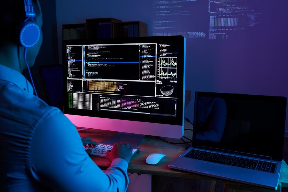 Programador trabalhando à noite em computador desktop com várias janelas de código aberto e laptop ao lado em ambiente escuro.